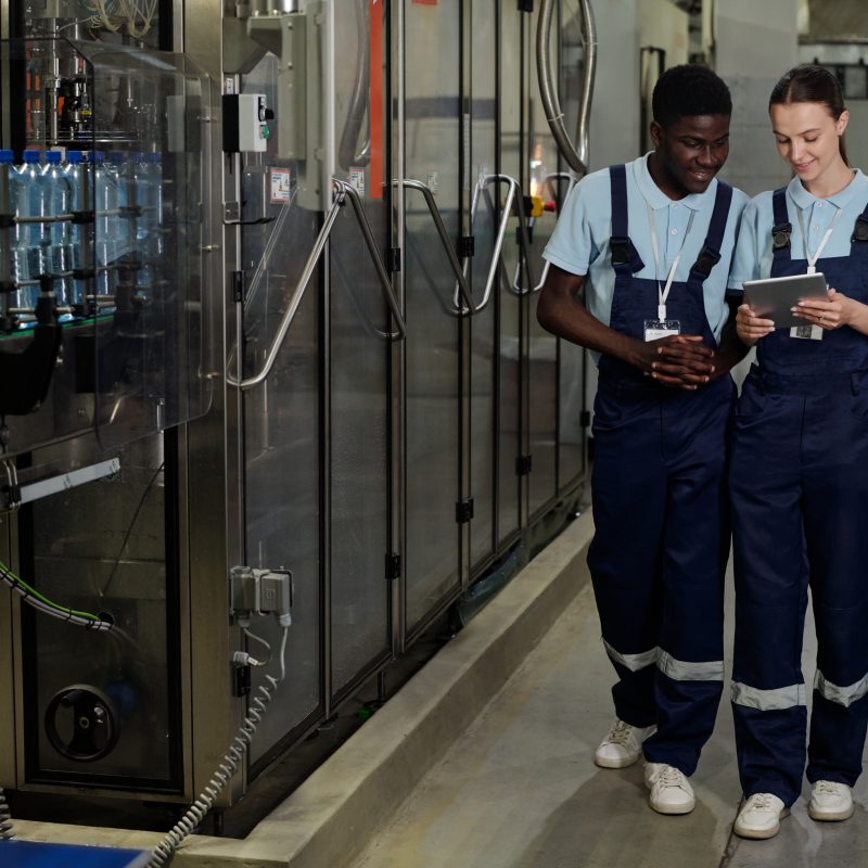 Young female and male engineers of quality control in workwear looking at screen of tablet while checking or discussing information